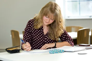 PhD student Sophie John concentrating while writing in a notepad on a desk in a student area