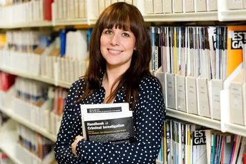Criminology PhD student Sophie Pike smiles are the camera while holding a textbook in front of bookshelves in the library