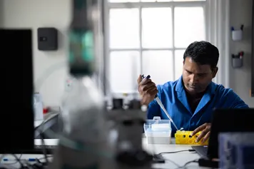 A student in blue overalls uses a pipet to transfer liquid into a test tube in a yellow test tube holder in a laboratory surrounded by scientific equipment