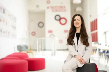 PhD student Gabrielle Hale smiles at the camera while sat in the foyer of the South Wales Business School at the Treforest campus with red sofas in the background