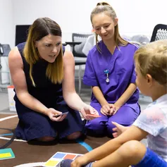 A nurse and a student nurse talk to a small child with picture cards and toys on the carpet of a paediatrician's office