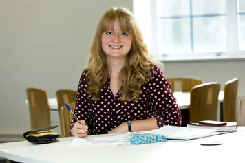 Sophie John, PhD student in Health Sciences and Psychology, sat at a table surrounded by pieces of paper, smiling at camera
