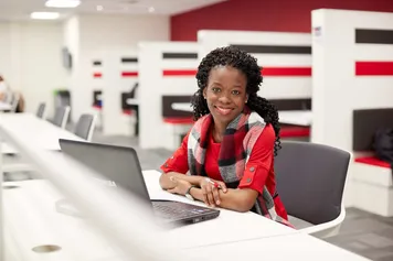 A student sat in the library working on a laptop and smiling at the camera.