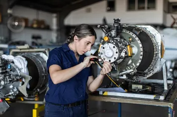 A person in a blue shirt uses a tool to work on a large mechanical component in an indoor industrial setting.