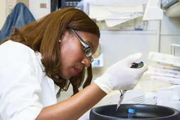 A student in a white laboratory coat, protective glasses and white rubber gloves uses a pipet to complete an experiment in a science lab