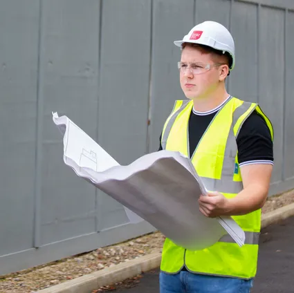 A quantity and surveying student wearing a yellow high visibility vest and a white USW hard hat overlooks a project while holding large white papers