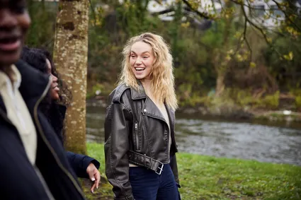 A student smiles while walking with their friends next to the grassy riverbank of the river Taff in Pontypridd