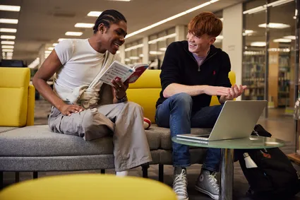 Two students looking at a book and using a laptop in the library.