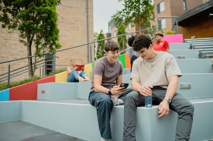 Students are sat on colourful steps, looking at a phone, outside the accommodation hub.