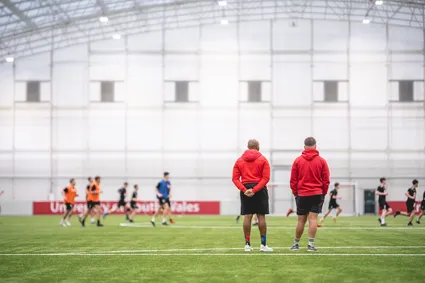 Students playing a game of football on the indoor 3g pitch. Two people are stood on the side line.