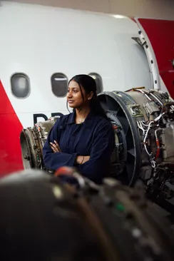 Student Pooja Umashankar poses in navy overalls in front of a jet enginge and a small aircraft