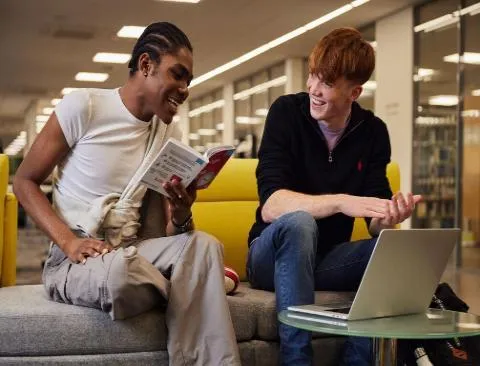 Two students looking at a book and using a laptop in the library.