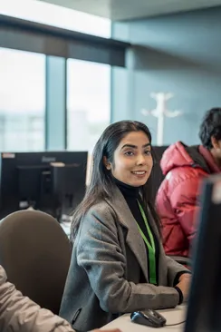 A cyber security academic wearing a grey coat and a green lanyard talks to a student off camera while working at a desktop computer with a student in a red puffer jacket and large windows behind themin a cyber security workshop