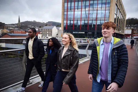 Four students walk together in a row on a footbridge over the river Taff in Pontyrpidd.