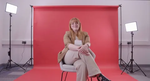university staff member sitting on a chair in front of a red background smiling to camera