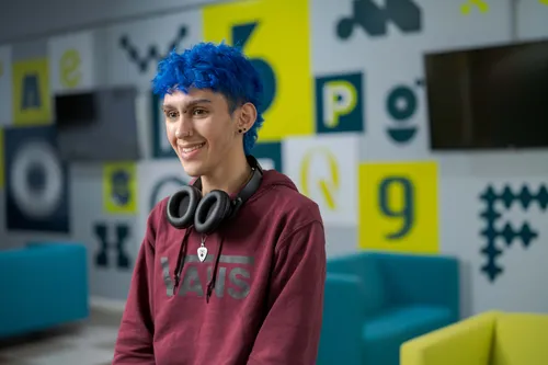 A person with short bright blue hair smiles indoors, wearing a maroon Vans hoodie and black headphones. The background is a modern, colorful space with teal seating and graphic wall designs.