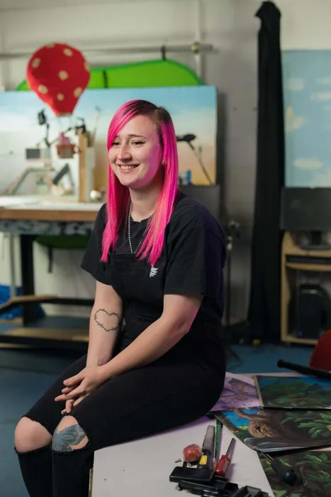 A profile shot of a woman with pink hair sitting at a table surrounded by various art supplies.