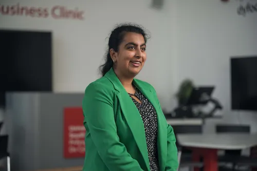 A smiling business student in a green blazer stands in front of a computer in a classroom setting.