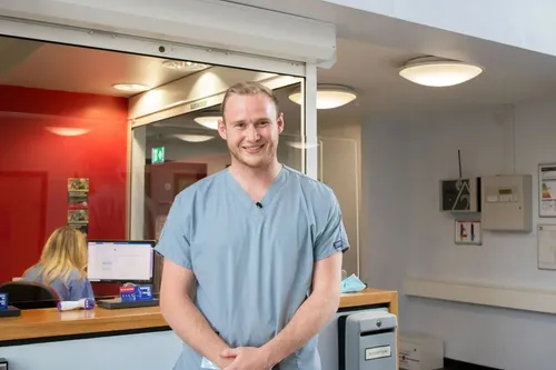 A chiro student in scrubs stands beside a reception desk, captured in a profile shot at a clinic.