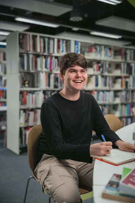 Smiling history student seated in a library, writing in a notebook at a table, profile shot with bookshelves in the background.