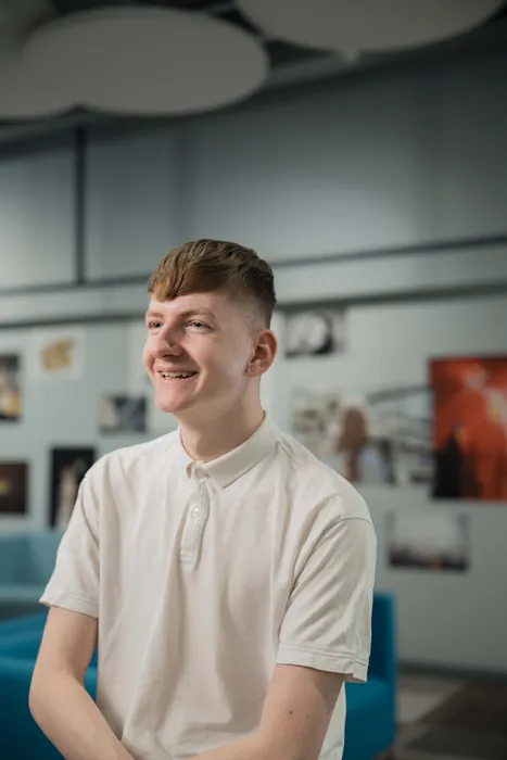 Marketing student, Sam, sits in front of colourful artwork whilst smiling at the camera