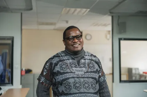A man wearing glasses and a patterned sweater over a turtleneck stands smiling in a well-lit room with desks and windows.