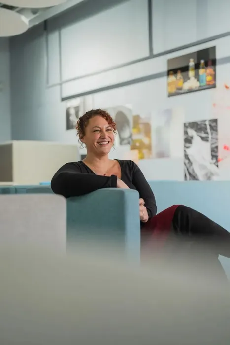 Photography student smiling for a photo, sitting on a sofa surrounded by a wall full of photographs