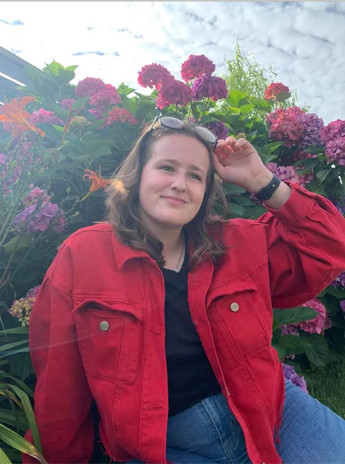 Postgraduate student Hannah Erpels sat amongst flowers and plants in a garden, smiling while holding sunglasses on their head
