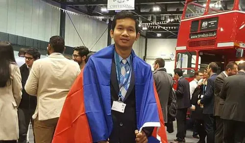 Student Koemhong Sol smiles at the camera while wrapped in a Cambodian flag in a transport museum with a lot of people chatting in the background