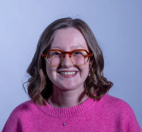 Interior Design student smiles at the camera while wearing a hot pink jumper and red glasses in front of a blue backdrop