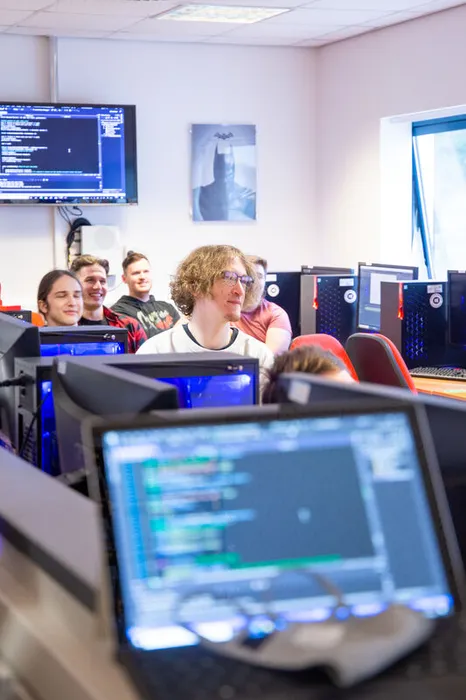A class of computing students working in a classroom. In the foreground there is a blurred laptop screen with coding on it.