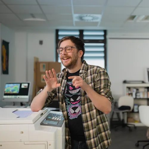 Illustration student, James, is leaning his elbow on a printer whilst talking and gesturing.