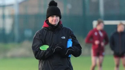 A football graduate in a black jacket and hat trains players, holding a green cone on the pitch.