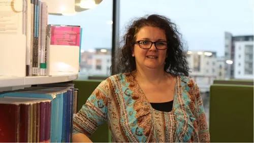 Teaching student, Julia, smiles at the camera whilst leaning on a bookshelf in Newport campus