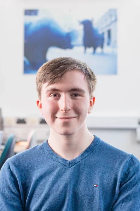 Studet Leo Humphreys smiles at the camera while wearing a blue t-shirt and sat in a computer room with various desktops on desks behind him