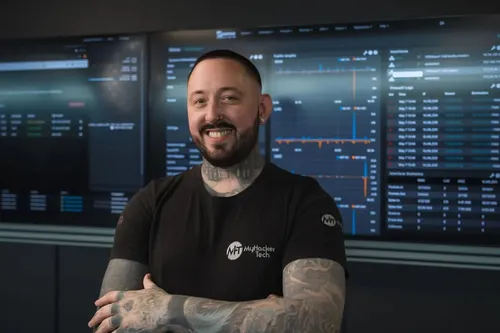 A smiling man with tattoos and a trimmed beard stands confidently with arms crossed in front of large digital screens displaying cybersecurity data and graphs. He is wearing a black 