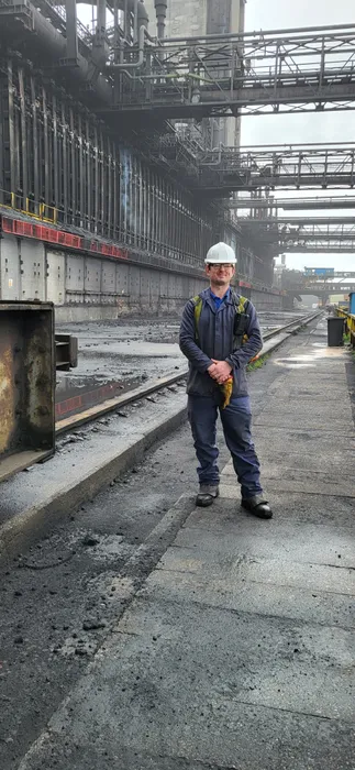 Engineering graduate Wayne stand proudly at his workplace wearing blue overalls and a hard hat