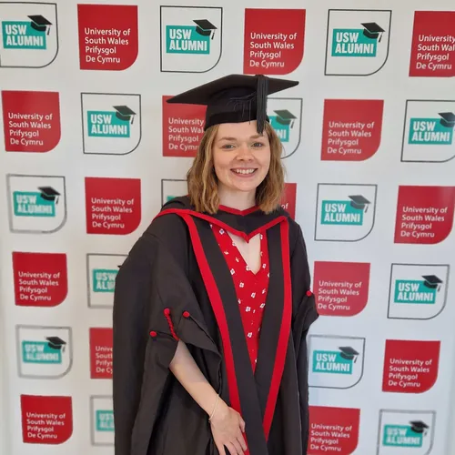 Ellie Rose is stood in front of a USW Alumni backdrop, wearing a graduation cap and gown.