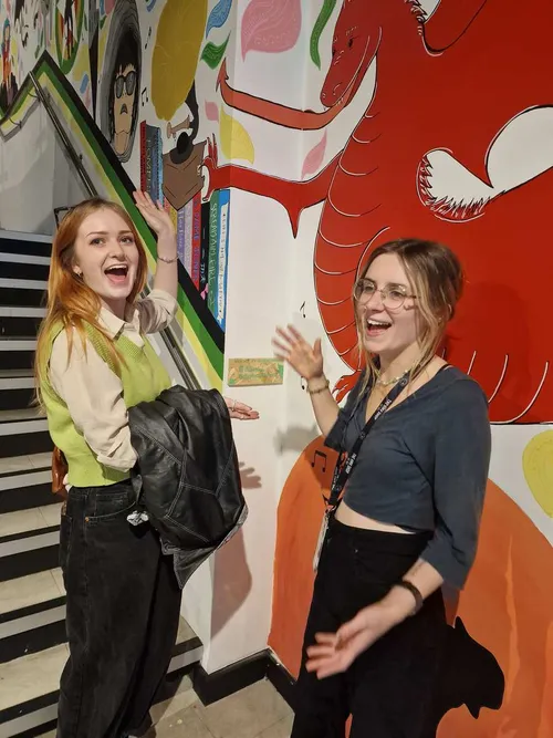 Graduates Sophie and Dali, smile at camera whilst pointing at some artwork