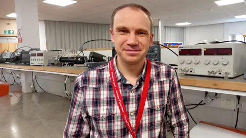 Darren Haines yn sefyll mewn labordy o flaen nifer o ddarnau o offer electronig