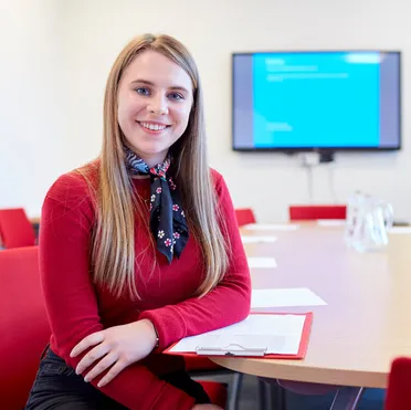 A student smiles at the camera while sitting down and leaning on a table in a meeting room at the Treforest Postgraduate Centre
