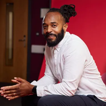 An international business academic smiles at the camera side on while sat in front of a red backdrop and leaning on their knees with their hands together