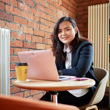 A smartly dressed student sat at their laptop with a coffee smiles at the camera from Crawshays Cafe in Treforest