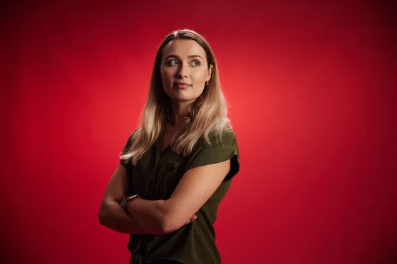 Alumni, Tara Peters, wearing a green shirt with her arms crossed stood in front of red backdrop looking in to the distance