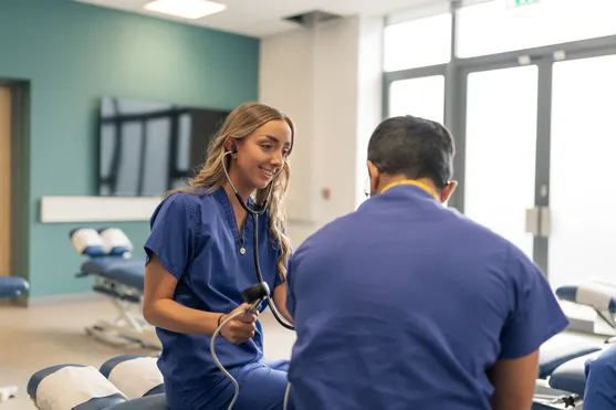 female student sat down with stethoscope, testing male student who has his back to the camera