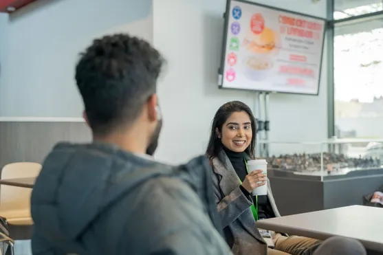 A cyber security student smiles at their friend and chats while drinking a coffee at Newport Campus