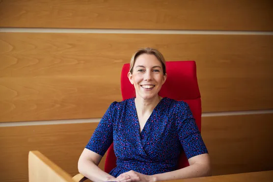An academic is sat in a tall red chair against a wooden wall. They are smiling into the camera.