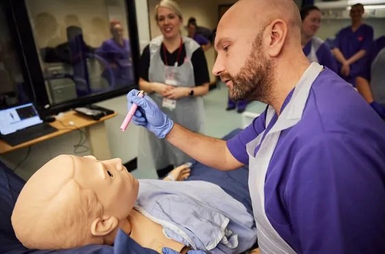 A nursing student is being supervised while carrying out an eye examination on an adult dummy.