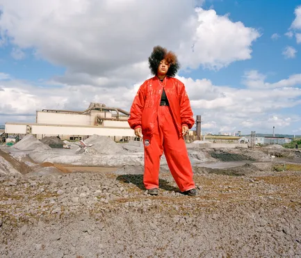 A person in a red suite, they are standing in front of a factory in a quarry.