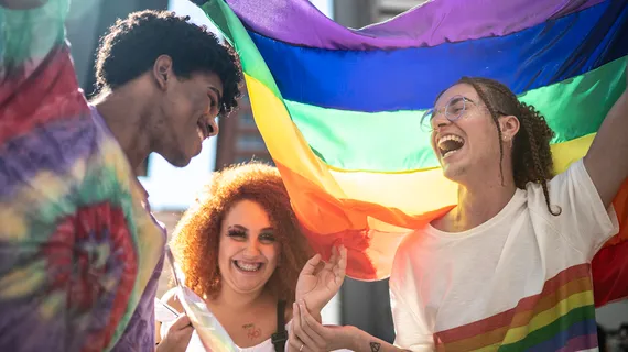 A group of three young adults at an LGBTQ+ event with a rainbow Pride flag